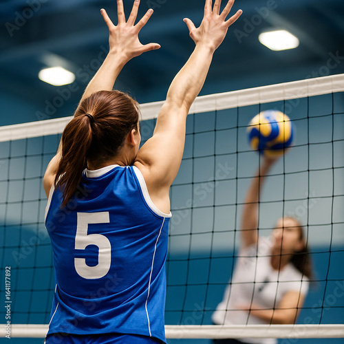 A female volleyball player in a blue jersey with number 5 jumps at the net to block a spike from an opponent during an indoor match.