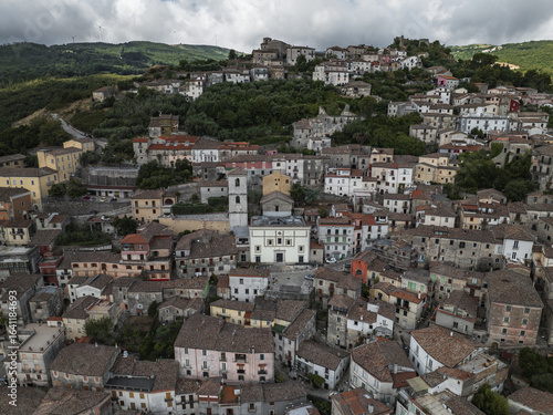 Aerial view of sun-kissed rooftops and winding streets cascade down the hillside, anchored by the church's stoic presence, Morcone, Benevento, Italy.