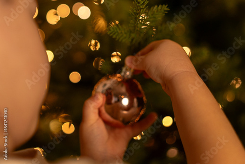 Child hanging ornament on Christmas tree with warm lights in festive setting
