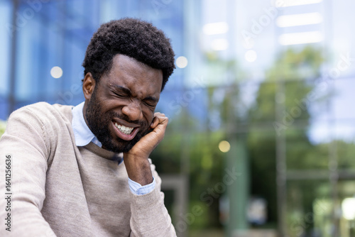 Tableau sur toile A young man sits outside a glass building, visibly wincing in pain, clutching his jaw clearly experiencing dental discomfort due to a toothache