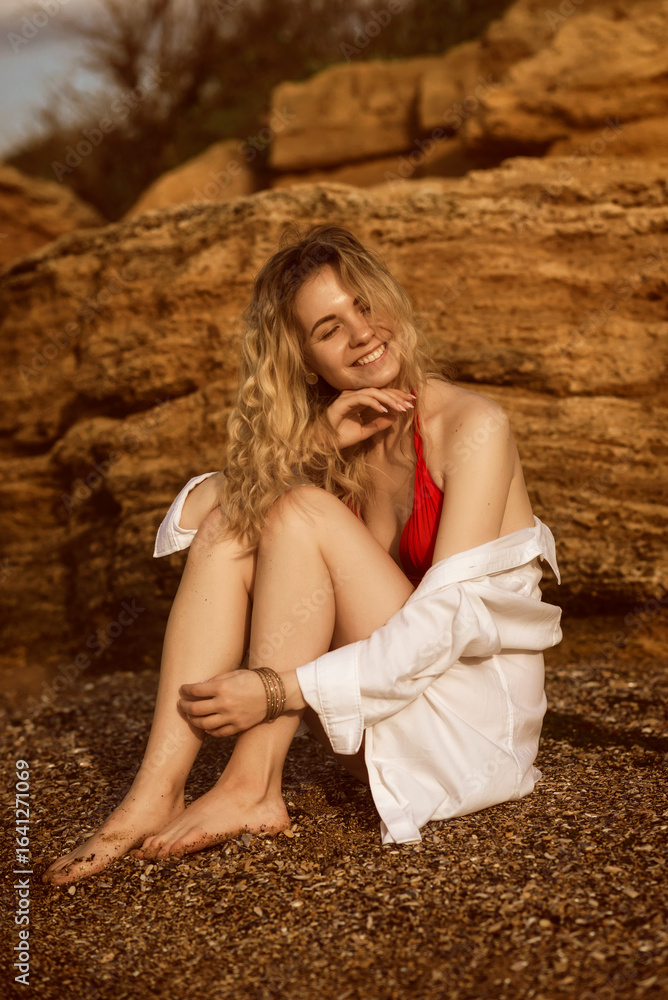 Naklejka premium Cute blonde in red swimsuit and white shirt sits on rock against sea background.