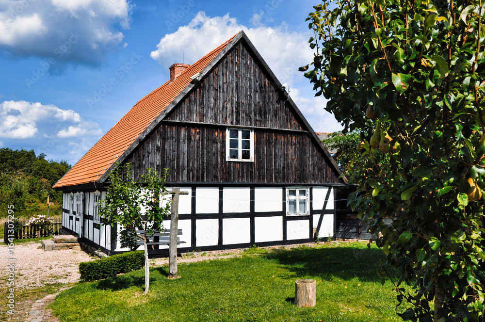 Fototapeta premium Swołowo - Malowniczy, tradycyjny dom z muru pruskiego otoczony bujnym ogrodem, na tle błękitnego, słonecznego nieba. Spokojna i urzekająca wiejska sceneria, podkreślająca rustykalny urok i piękno.