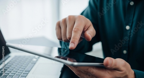 A close-up of a man's finger pointing at and interacting with the screen of a tablet computer, with a laptop blurred in the background.