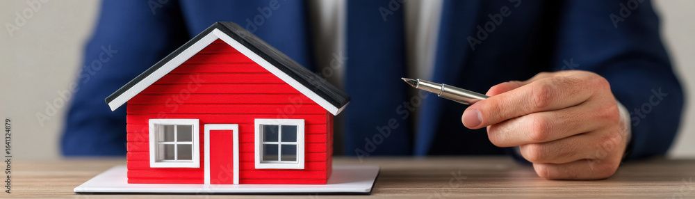 Businessman holding pen near small red house model on wooden table, symbolizing real estate investment and property deal Businessman holding pen near small red house model on wooden table, symbolizing real estate investment and property deal