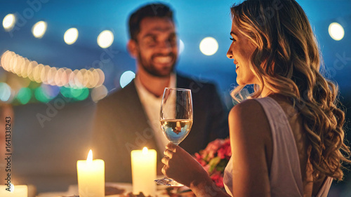 A happy couple on a romantic outdoor dinner date, with the woman holding a glass of white wine. The scene is lit by soft, glowing candles and string lights, creating a warm and intimate atmosphere.