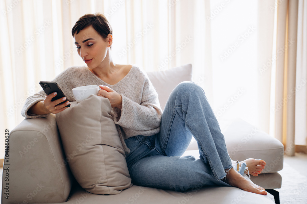Obraz premium Woman sitting on sofa , drinking tea and using mobile phone