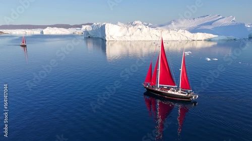 Little red sailboat cruising among floating icebergs in Disko Bay glacier during midnight sun season of polar summer. Ilulissat, Greenland. Global warming and melting glaciers.