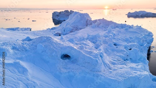 A tall melting iceberg in the Arctic Ocean. A glacier with a lake and waterfall. Clear ice floats in turquoise water. Ecology, melting ice, climate change, global warming. Aerial view. Midnight sun.