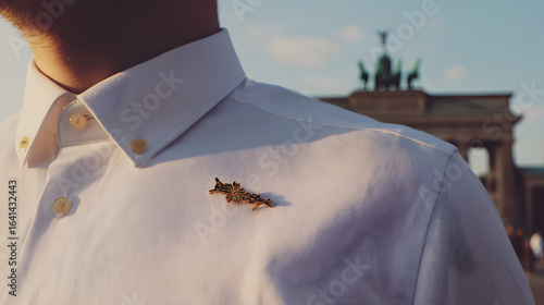 White shirt with black-red-gold lapel pin, shallow focus on blurred Brandenburg Gate in morning light.