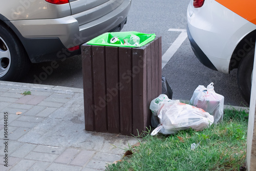 There is a full trash can on the sidewalk and bags of trash nearby