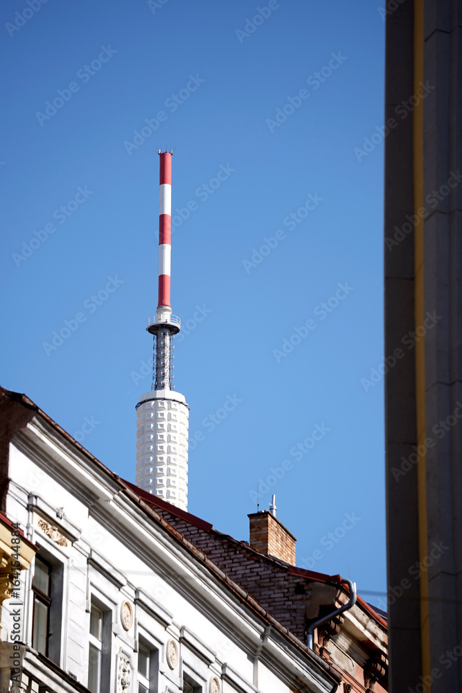 Obraz premium Tall communication tower with red and white stripes rises above rooftops against a clear blue sky. Architectural details of the building roof and chimney are visible. Clear daytime urban scene.