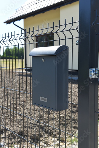 Contemporary mailbox attached to black steel mesh fence, in front of new residential house.