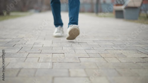 Person walking on paved sidewalk in city park, captured from low angle with motion blur on legs and shoes, emphasizing movement, journey, and passage of time in peaceful outdoor environment