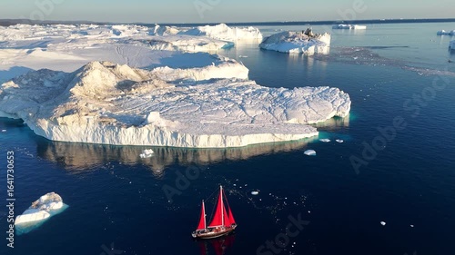 Little red sailboat cruising among floating icebergs in Disko Bay glacier during midnight sun season of polar summer. Ilulissat, Greenland. Global warming and melting glaciers.