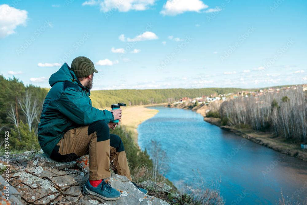 Naklejka premium A man sits on a rock overlooking a river, holding a cup of coffee