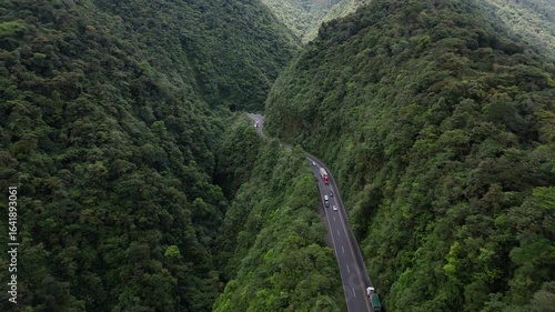 Aerial push forward along mountain road through lush forest with distant tunnel in the background