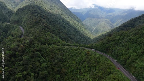 Aerial drone push forward over winding mountain road through lush green valley with cars and trucks driving