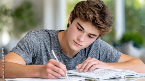 A teenager is focused on his homework, writing notes in an open notebook while sitting at a table with books nearby