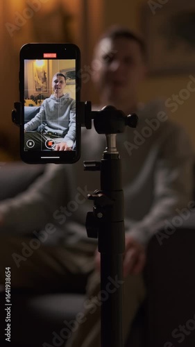Male blogger recording motivational video at home in cozy evening atmosphere with warm lighting, smiling and positive, content creator lifestyle, vertical video
