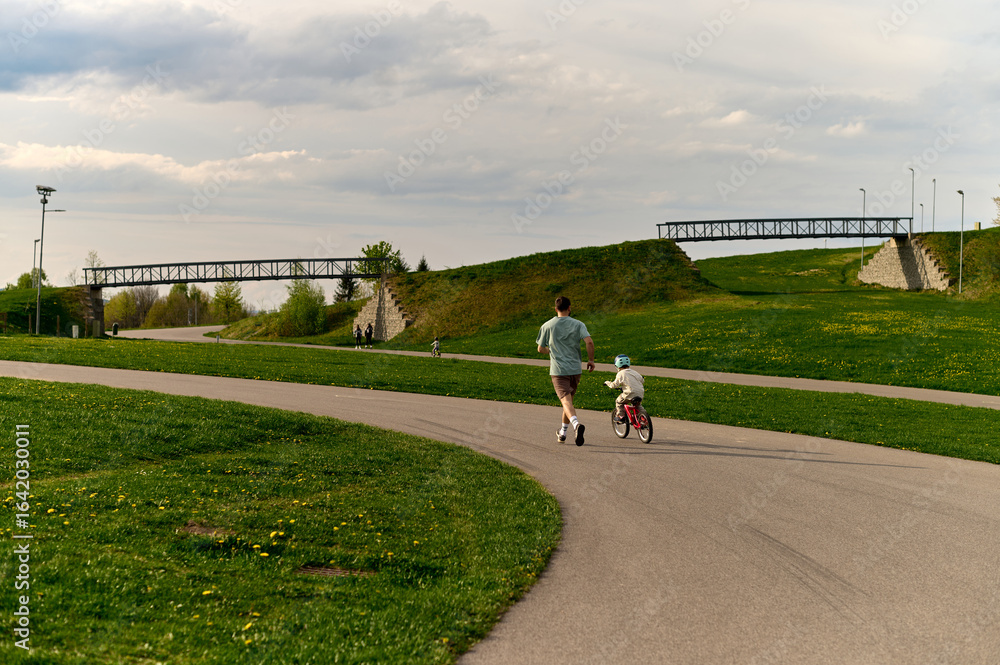 A father jogs next to his young child riding a red bicycle A father jogs next to his young child riding a red bicycle
