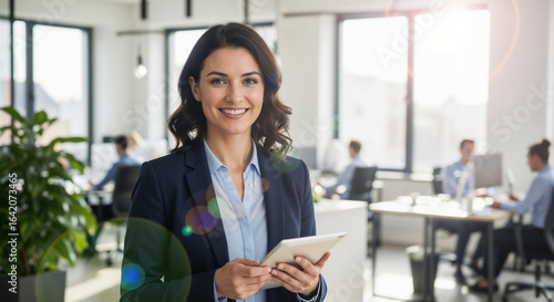 Professional woman in formal attire holding a tablet in a contemporary office setting with engaged coworkers. Ideal image for corporate, teamwork, and professional themes.

