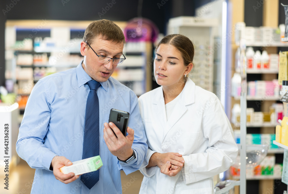 Obraz premium Concerned middle-aged man showing to young female pharmacist QR-code of product on his smartphone in chemist's shop