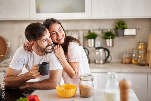 A couple shares joyful moments in a bright kitchen while preparing breakfast. The man holds a coffee cup, and the woman hugs him, smiling happily amidst various ingredients.