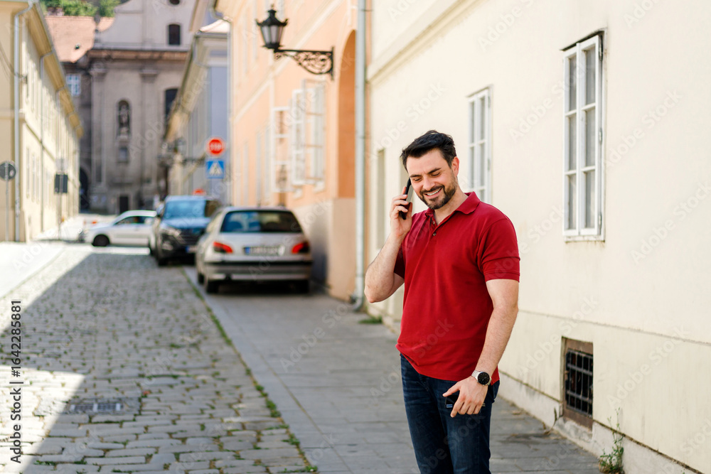 Fototapeta premium Man smiles while talking on phone in a charming cobblestone street during the day