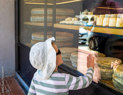 A toddler wearing a sun hat and striped shirt gazes with curiosity at a cheese shop display in Le Touquet, France, pressing their hand against the glass in fascination.