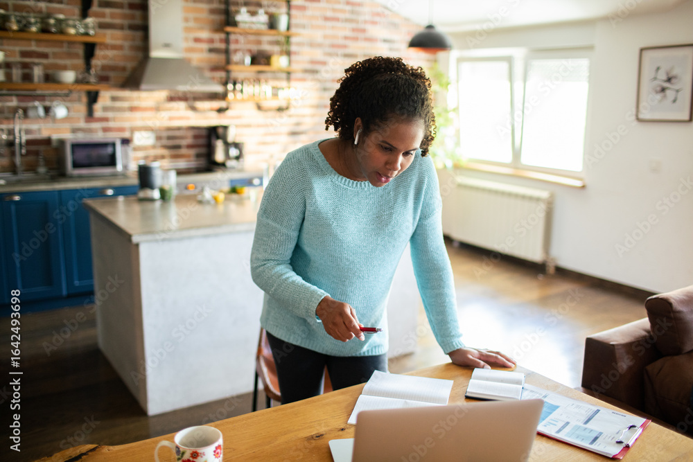 Black woman working from home kitchen on video call with laptop and financial documents on table Black woman working from home kitchen on video call with laptop and financial documents on table