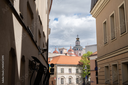 narrow urban alleyway with ornate historic church towers under a partly cloudy sky, framed by residential buildings, displaying a juxtaposition of old and modern architecture, sunlit, serene