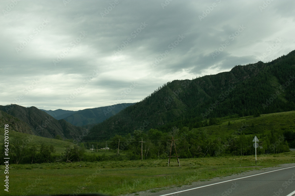 Naklejka premium Wide view of a rural landscape of Altai under dramatic storm clouds, with green hills and mountains in the distance, crossed by power lines. Russian nature.