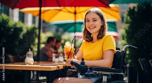 A smiling young woman in a wheelchair enjoys a cold drink at an outdoor cafe on a sunny day.