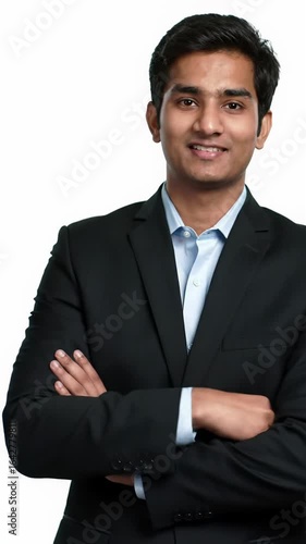 Young indian man in a dark business suit and light blue shirt, confidently standing with crossed arms, smiling at camera in a professional studio with a pristine white background and slow zoom-in,