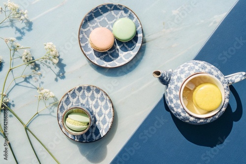 French Dessert Afternoon Tea with flat lay of macarons, lemon tart, and a small teapot with tea cups on a marble table with delicate flowers.