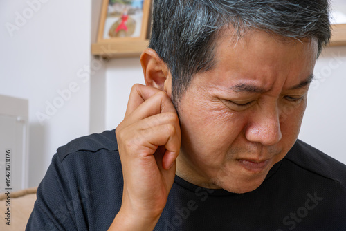 An Asian man clutching their ear in visible discomfort, set against a home interior—ideal for medical ads on ear pain, hearing loss, and neurological treatment.