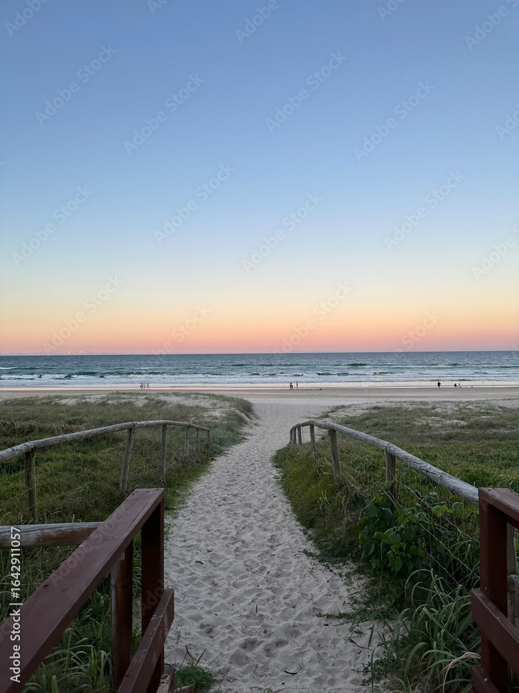 Obraz premium Wooden pathway leading through soft white sand dunes towards the open ocean at sunset, symbolizing peace, escape, and the beauty of untouched coastal landscapes.