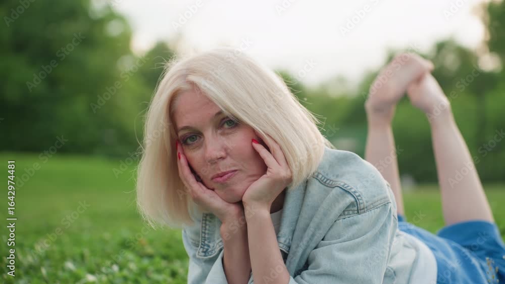 stylish woman lying on green grass in park resting chin on hands, thoughtful gaze, casual denim jacket, soft background of blurred trees and warm light, relaxed mood conveying serenity stylish woman lying on green grass in park resting chin on hands, thoughtful gaze, casual denim jacket, soft background of blurred trees and warm light, relaxed mood conveying serenity
