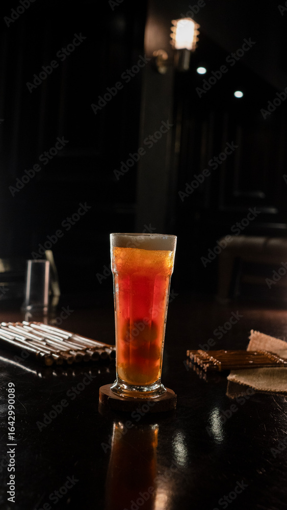 A tall glass of iced lychee tea with a vibrant orange gradient, containing whole lychee fruits at the base, served on a wooden coaster over a dark reflective surface, creating a refreshing and tropica A tall glass of iced lychee tea with a vibrant orange gradient, containing whole lychee fruits at the base, served on a wooden coaster over a dark reflective surface, creating a refreshing and tropica