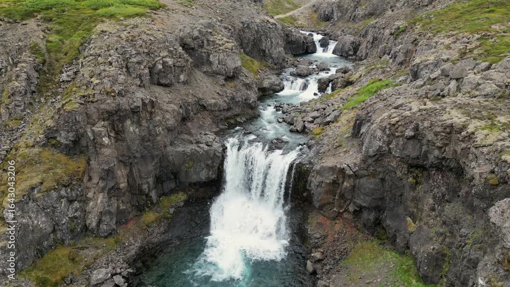 Aerial view of a powerful waterfall cascading into a clear pool, framed by rugged cliffs with patches of green moss, creating a dramatic contrast, Egilsstaðir, Múlaþing, Iceland. Aerial view of a powerful waterfall cascading into a clear pool, framed by rugged cliffs with patches of green moss, creating a dramatic contrast, Egilsstaðir, Múlaþing, Iceland.