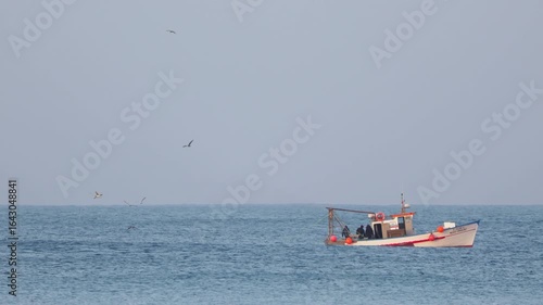 Fishermen at sea on a fishing boat, loading crates of freshly caught fish while seagulls fly around. Clear sky and calm weather.