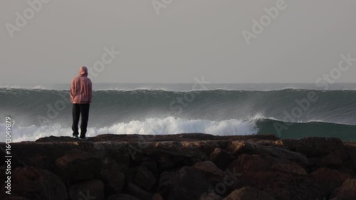 Back view of a man standing on a stone pier at Costa da Caparica, Portugal, admiring a powerful morning wave under a clear yet cold sky.