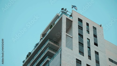 A part of a residential building, in a low angle telephoto view, where a single Israeli flag is hanged out from the top of the apartment building, flapping lightly with the wind. Ramat Gan, Israel.