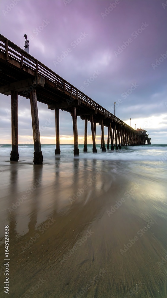Obraz premium Long wooden pier extending into ocean at sunset, reflecting in wet sand