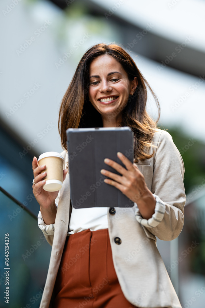 Fototapeta premium Smiling businesswoman using digital tablet and holding coffee outside office building