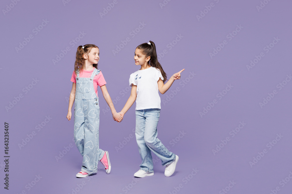Naklejka premium Full body smiling fun little two friends kids children girls 9-10 years old wear casual clothes together hold hands walk go point finger aside isolated on plain purple background. Lifestyle concept.