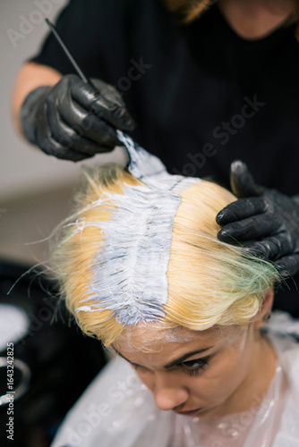 A professional hairstylist applies hair color to a female client's blonde hair in a salon. The stylist, wearing black gloves, carefully works on the roots.