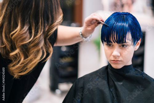 A female hairdresser combs the blue hair of a young female client in a salon. The client wears a black cape with blue hair clippings, indicating a recent haircut or styling session.