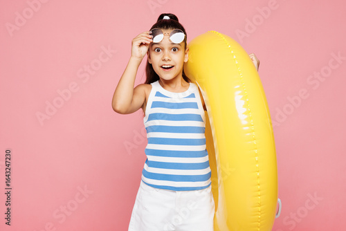 Фотография Traveler happy kid child wearing blue casual clothes rest near hotel pool hold inflatable rubber yellow ring take off sunglasses isolated on plain pink background