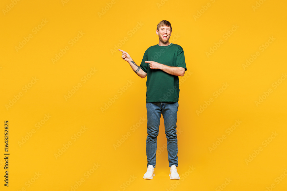 Fototapeta premium Full body young smiling happy man wear green t-shirt casual clothes point index finger aside on blank empty area mock up isolated on plain yellow orange background studio portrait. Lifestyle concept.